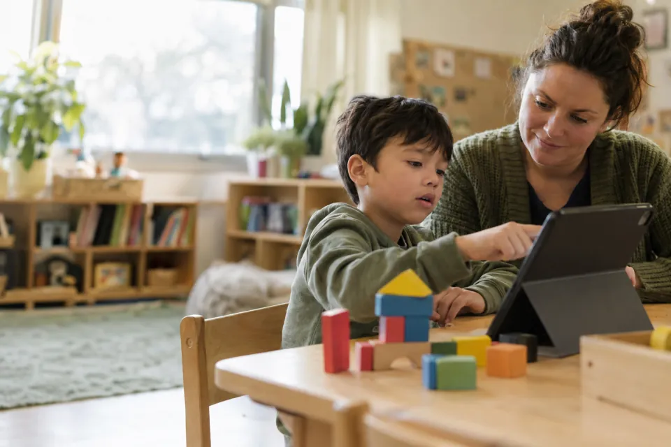 A student uses a tablet beside an adult at a classroom table with colorful building blocks.